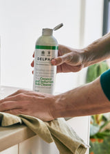 A person in a teal shirt cleans a dark countertop with a cloth while holding a Delphis cleaning spray bottle. A matching Delphis cleaner bottle and a metal colander with leafy greens sit on the counter in a bright kitchen setting.